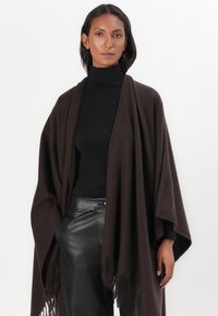 Brauner Wollponcho mit Fransenverzierungen, der ein lockeres Design und eine weiche Textur aufweist, getragen über einem schwarzen Rollkragenpullover und Lederhosen.
