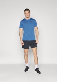 Blaues, kurzärmeliges Sportshirt mit strukturiertem Design, kombiniert mit schwarzen Shorts. Model trägt schwarze Turnschuhe und weiße Socken.