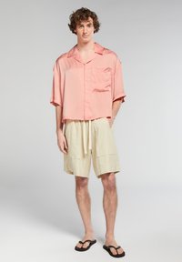Camicia in satin rosa chiaro a maniche corte con tasca, abbinata a pantaloni corti in lino beige con coulisse, e ciabatte nere.