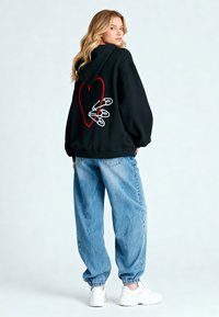 Felpa nera con un design a forma di cuore rosso sul retro, decorata con spille da balia. Abbinata a jeans larghi blu chiaro e sneakers bianche.