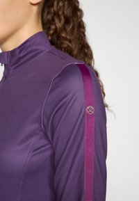 Vijolična dolgosleeve atletska majica z visokim ovratnikom, teksturirano blago, kontrastne magenta stranske črte in logotipom na rokavu.