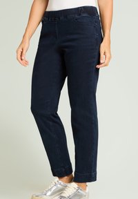 Donna che indossa jeans slim-fit blu scuro con vita elastica e sneakers argento, in piedi su uno sfondo chiaro e semplice.