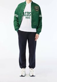 Man wearing green Lacoste jacket, white Lacoste Paris polo, navy track pants, and white Lacoste sneakers on white background.