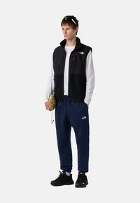Chaleco negro con acentos de forro polar, camiseta blanca de manga larga, pantalones de chándal azul marino y zapatillas negras. Incluye una pequeña bolsa beige.