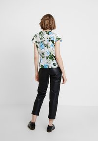 Blusa a maniche corte con motivo floreale bianco, caratterizzata da foglie verdi e blu. Abbinata a pantaloni in pelle neri e aderenti e scarpe nere.