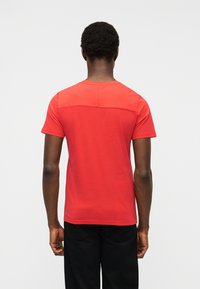 Άνδρας με κοντά σγουρά μαλλιά, όρθιος με την πλάτη προς το μέρος μας, φοράει κόκκινη κοντομάνικη μπλούζα και μαύρο παντελόνι μπροστά σε απλό φόντο.