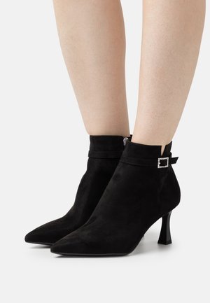 Bottines en suède noir avec des pointes effilées, un petit talon carré et un détail de boucle décoratif au niveau de la cheville. Texture lisse et design épuré.