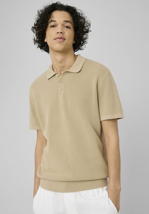 Jeune homme aux cheveux bouclés portant un polo beige texturé et un pantalon blanc, debout devant un fond gris clair.
