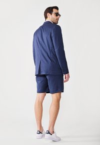 Veste de costume bleu marine avec une seule fente, associée à un short assorti. Portée avec des baskets blanches et de lunettes de soleil sombres.