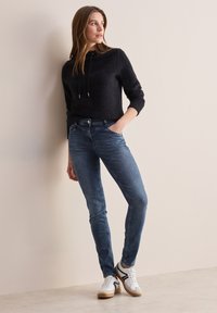 Suéter negro recortado con cordón, jeans ajustados azules y zapatillas blancas con acentos en gris y negro, todo contra un fondo neutro.