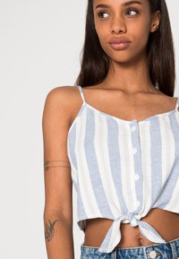 Top cropped em azul e branco com riscas, com fecho de botão na frente e detalhe de laço na bainha. Feito de um tecido leve com alças finas.