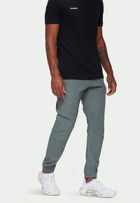 Moški v črni majici Mammut, sivih športnih hlačah in belih športnih čevljih stoji ob enobarvni ozadju.