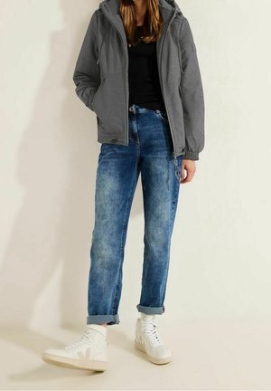 Persona que lleva una chaqueta gris con capucha, top negro, vaqueros azules remangados y zapatillas blancas de caña alta, de pie frente a un fondo claro y liso.