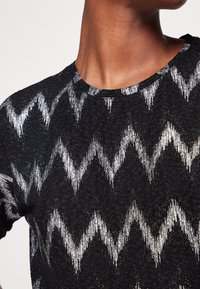 Närbild av en person som bär en svart tröja med silverfärgade zickzackmönster och rund halsringning.