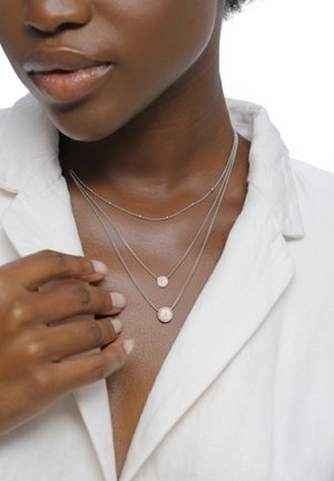 Drei geschichtete Silberketten: eine mit einem runden Anhänger mit irisierendem Muster, eine zweite mit einer strukturierten Scheibe und eine zarte Kette.