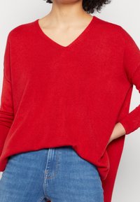 Czerwony sweter z dekoltem w serek i długimi rękawami, wykonany z miękkiego materiału, luźny krój i podkreślony dół; zestawiony z jasnoniebieskimi dżinsami.