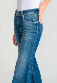 Jeans flare in denim blu con vita alta, tasche frontali e chiusura con bottone dorato, caratterizzati da un lavaggio chiaro e lievi segni di usura.
