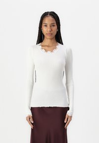 Valkoinen ribattu pitkähihainen paita, jossa on aaltoileva kaula-aukko ja pitsireunus. Sujuva rakenne, slim fit -malli ja istuvat hihansuut. Yhdistetty burgundinväriseen hameeseen.
