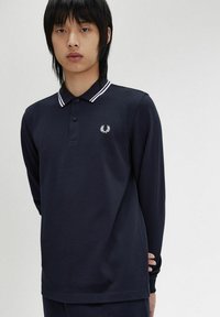 Polo a maniche lunghe blu navy con colletto a strisce bianche, pattina con tre bottoni e logo ricamato sul petto. Tessuto di cotone liscio.