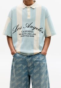 Modré a krémové pruhované polo tričko s texturovaným pleteným vzorem, vybavené tříknoflíkovou légou a černým potiskem textu. Nošeno s světle modrými džínami zdobenými opakujícím se vzorem loga.