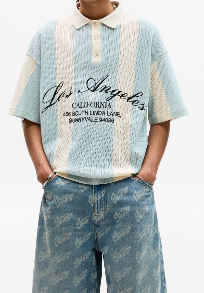 Modré a krémové pruhované polo tričko s texturovaným pleteným vzorem, vybavené tříknoflíkovou légou a černým potiskem textu. Nošeno s světle modrými džínami zdobenými opakujícím se vzorem loga.