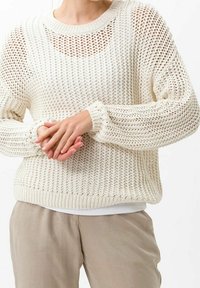 Pull en tricot couleur crème avec un tissage lâche, col rond et poignets côtelés, superposé sur une chemise blanche. Tissu léger et texturé.