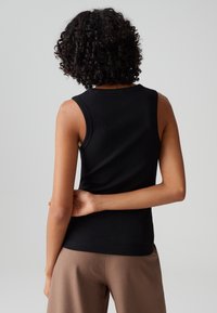 Personne aux cheveux bouclés portant un haut noir sans manches et un pantalon beige, debout avec une main dans le dos.