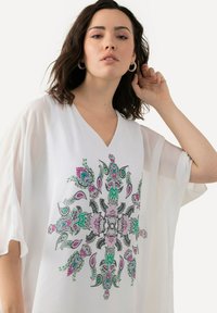 Tunic bianca con scollatura a V e maniche trasparenti, caratterizzata da un vivace motivo floreale e paisley nei colori verde, rosa e nero al centro.