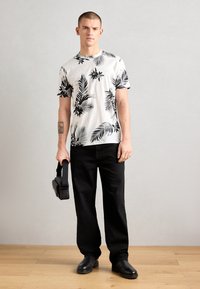 T-shirt met korte mouwen en ronde hals in het wit met zwart tropisch bladmotief, gecombineerd met zwarte broek en zwarte schoenen, en een handheld apparaat in de hand.