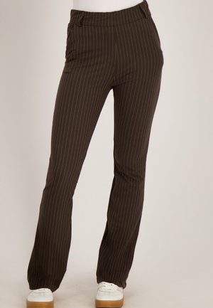 Bruine rechte broek met krijtstreep, gedragen met witte sneakers door een persoon die staat tegen een effen achtergrond.