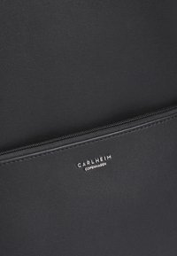 Črna sintetična usnjena torba z gladko teksturo, z žepom na zadrgo in vtisnjenim logotipom "CARLHEIM COPENHAGEN" v srebrni barvi.