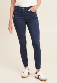 Donkerblauwe skinny jeans met een middelhoge taille, voorzakken en een knoopsluiting, gecombineerd met witte sneakers met zwarte accenten.