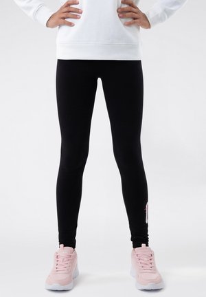 Persona in piedi con le mani sui fianchi, indossa leggings neri e scarpe da ginnastica rosa su uno sfondo bianco neutro.