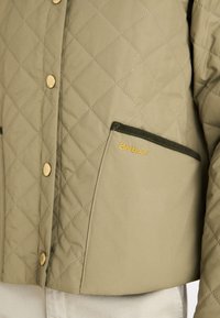 Quilted olive green jacket with gold-tone snap buttons and a Barbour logo. Features a textured finish and chest pocket with green trim.