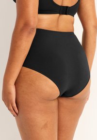 Kvinna som bär högmidjade svarta trosor och en matchande svart bh, sedd bakifrån, mot en enfärgad ljus bakgrund.