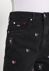 Jeans de mezclilla negra con bordados de corazones en rojo, blanco y azul. Presenta un diseño clásico de cinco bolsillos y cierre de botón.