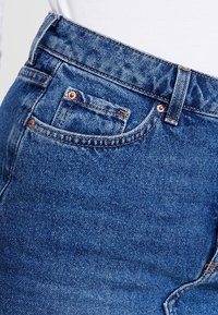 Jeans in denim blu medio con una texture liscia, caratterizzati da accessori in rame, una tasca frontale e un design a vita alta.