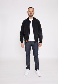 Svart bomberjacka med dragkedja fram, sidofickor och ett märke på vänstra nedre delen. Bärs över en vit skjorta och mörka jeans med vita sneakers.