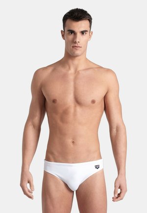 Giovane uomo in piedi di fronte, indossa slip da bagno bianchi con un piccolo logo nero sull'anca sinistra su uno sfondo chiaro e uniforme.