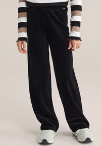 Čierne kordúrové nohavice s širokými nohavicami a ribbovanou textúrou, s uvoľneným strihom a elastickým pásom. Skombinované s svetlými teniskami.