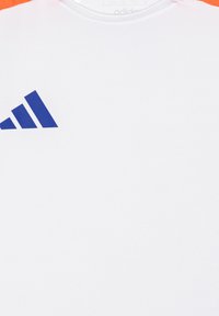 Camiseta atlética blanca con el logo azul de Adidas en el lado izquierdo del pecho, y detalles de tela naranja en los hombros. Textura suave, escote redondo.