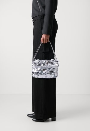 Osoba ubrana na czarno trzymająca srebrną torebkę pokrytą cekinami z łańcuszkowym uchwytem na jednolitym białym tle.
