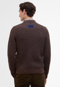 Brown ribbed knit sweater featuring a high collar and a blue square patch on the back. The fit is relaxed, with a textured surface.