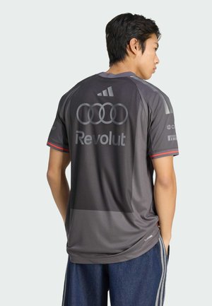 Man die van achteren kijkt en een donkergrijs sportshirt draagt met Audi- en Revolut-logo's, gecombineerd met blauwe broek met witte strepen.