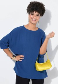 Top blu, oversized e testurizzato con scollatura rotonda e maniche a tre quarti. Presenta una borsa gialla e dettagli in gioielli dorati.