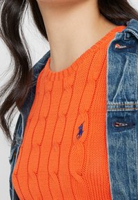 Zbliżenie na osobę w pomarańczowym swetrze w warkocze z niebieskim haftowanym logo i niebieskiej dżinsowej kurtce zarzuconej na ramiona.