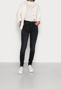 Chaquetón de mezclilla blanca sobre una blusa clara, combinado con jeans skinny oscuros y zapatillas blancas. La chaqueta tiene detalles de botones y un dobladillo con estampado.
