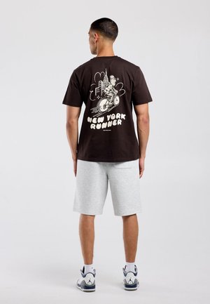 Man draagt een zwart T-shirt met "New York Runner" afbeelding van een fietser en stadsgezicht, lichtgrijze korte broek en witte sneakers, staand met de rug naar voren.