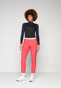 Black long-sleeve top with logo, paired with bright pink fitted pants and white belt. Model wears white athletic shoes and ankle socks.