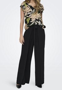 Pantalon noir à jambes larges avec une ceinture à nouer, associé à un chemisier à manches courtes arborant un imprimé floral tropical aux tons beige et vert.